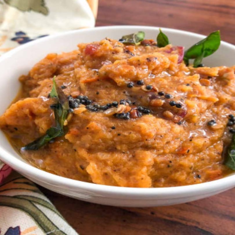 A white bowl filled with orange-brown Indian chutney, garnished with black seeds and green curry leaves.